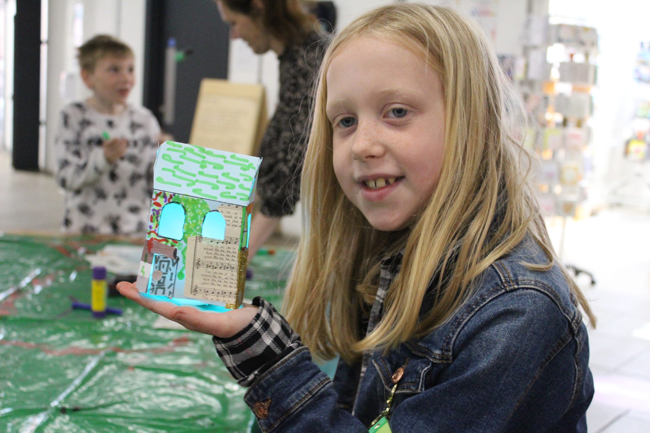 A tween girl holds up a light up house that she has made from card and decorated.