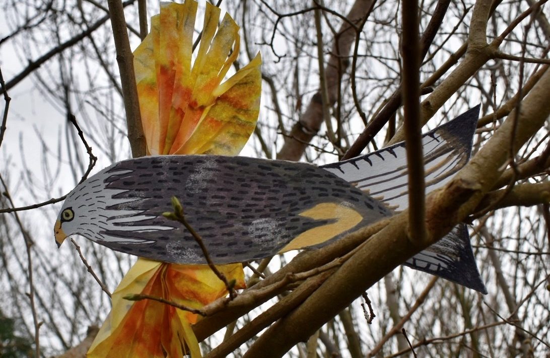 A bird of prey made from painted cardboard and recycled coffee filters perched in a tree outdoors.